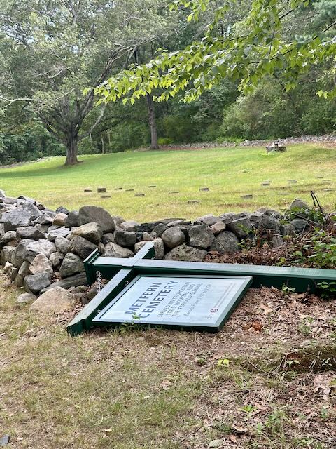 A sign reading "MetFern Cemetary" lays on its side.  Behind the sign, a knee-high stone wall marks a grassy field with low grave markers and a single tree.